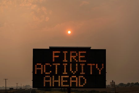 A highway sign reads, "FIRE ACTIVITY AHEAD," in the foreground, the landscape, sky, and sun in the background can be seen shrouded with heavy wildfire smoke, causing a warm, orange tint to be cast